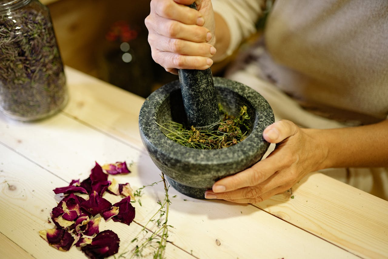 A person grinding herbal ingredients in a stone mortar and pestle with dried petals nearby.